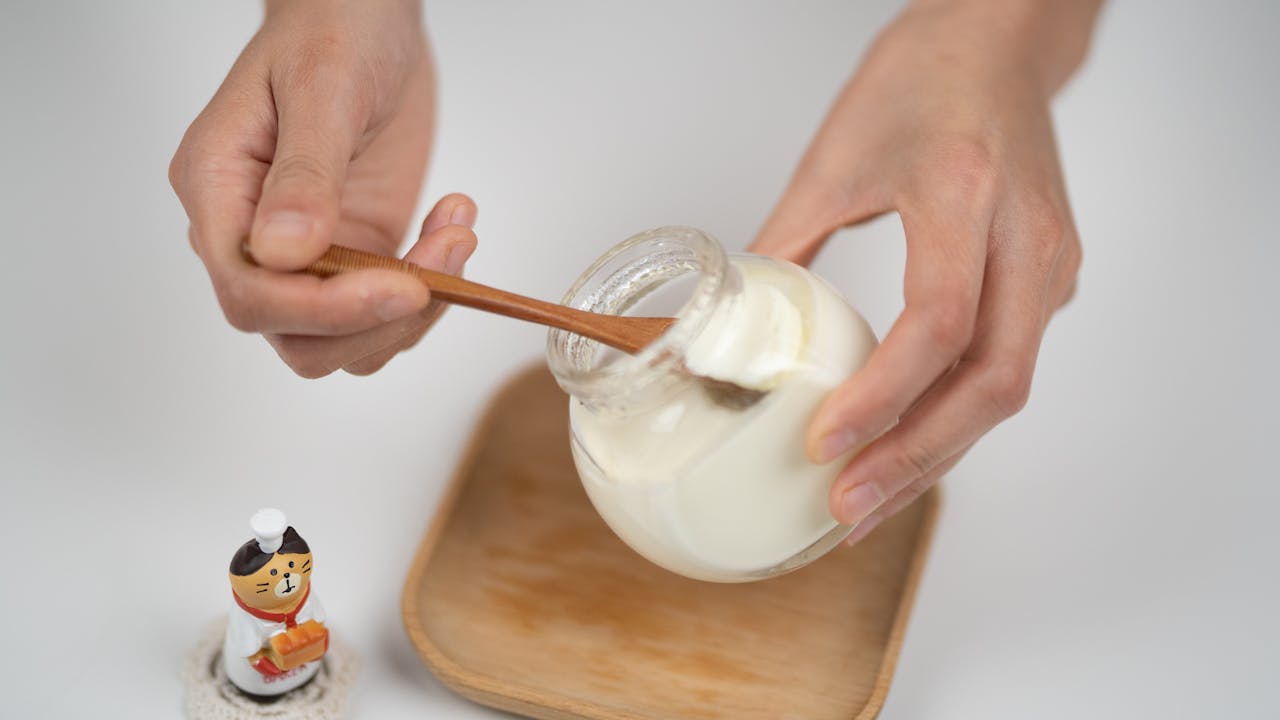 services-03 Close-up of hands serving creamy yogurt from a jar using a wooden spoon, with a cute figurine on the side.